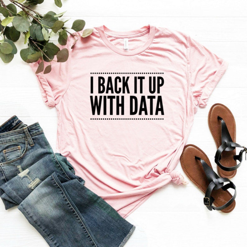 May include: A light pink short-sleeved t-shirt featuring the black text I BACK IT UP WITH DATA centered in a rectangular frame with dotted borders. The shirt is displayed alongside blue denim jeans and a pair of brown leather thong sandals.