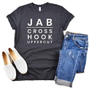May include: A dark gray t-shirt with white text that reads "JAB CROSS HOOK UPPERCUT". The shirt is paired with blue jeans and white slip-on shoes.