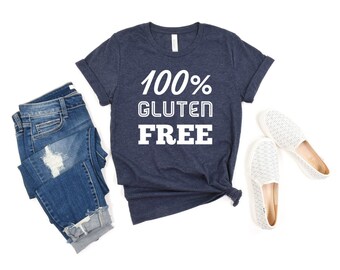 May include: A dark heather navy blue short-sleeve t-shirt with the white text 100% GLUTEN FREE printed on the front, paired with blue denim jeans featuring distressed details and a pair of white slip-on shoes with a textured fabric pattern.