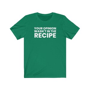 May include: A short-sleeved green t-shirt featuring the white text "YOUR OPINION WASN'T IN THE RECIPE" printed in a bold, sans-serif font across the centre of the chest. The garment has a crew neckline and a standard, straight-cut silhouette.