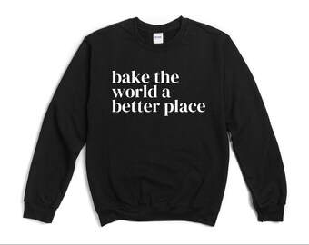 May include: A black long-sleeve crewneck sweatshirt featuring the white text "bake the world a better place" printed across the chest. The garment has a ribbed collar, cuffs, and hem, with a standard fit and a Gildan brand label at the neckline.