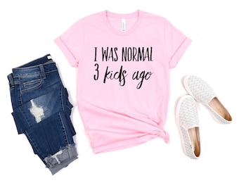 May include: A light pink short-sleeved t-shirt with the black text I WAS NORMAL 3 kids ago, paired with dark blue distressed denim jeans and a pair of white slip-on shoes with a textured pattern on the upper fabric surface.