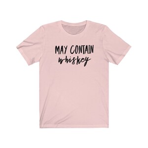 May include: A light pink t-shirt with the phrase "MAY CONTAIN whiskey" printed in black cursive lettering. The shirt has short sleeves and a crew neck. The fabric appears to be soft and comfortable.