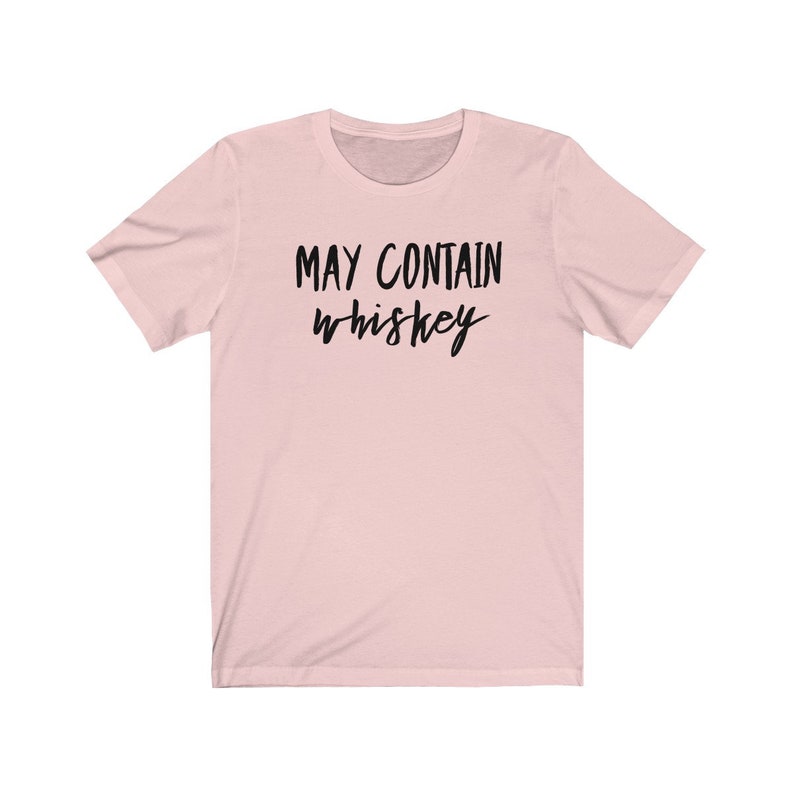 May include: A light pink t-shirt with the phrase "MAY CONTAIN whiskey" printed in black cursive lettering. The shirt has short sleeves and a crew neck. The fabric appears to be soft and comfortable.