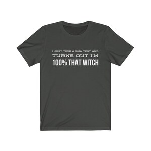 May include: A dark gray short-sleeved t-shirt featuring white text printed on the chest that reads, I JUST TOOK A DNA TEST AND TURNS OUT I'M 100% THAT WITCH. The garment has a crew neckline and a standard fit, displaying clear, bold lettering.