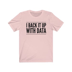 May include: A light pink short-sleeved t-shirt featuring the black text "I BACK IT UP WITH DATA" centered on the chest, enclosed between two horizontal lines of small dots above and below the lettering, with a classic crew neckline.