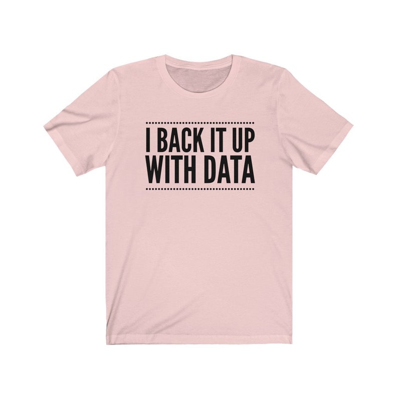 May include: A light pink short-sleeved t-shirt featuring the black text "I BACK IT UP WITH DATA" centered on the chest, enclosed between two horizontal lines of small dots above and below the lettering, with a classic crew neckline.