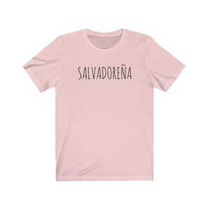 以下が含まれることがあります： 前面に黒い文字で「SALVADORE&Ntilde;A」とプリントされたピンクのTシャツです。