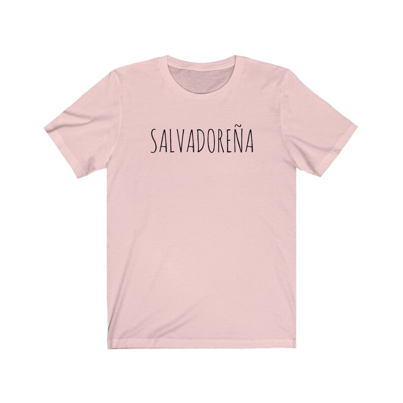 以下が含まれることがあります： 前面に黒い文字で「SALVADORE&Ntilde;A」とプリントされたピンクのTシャツです。