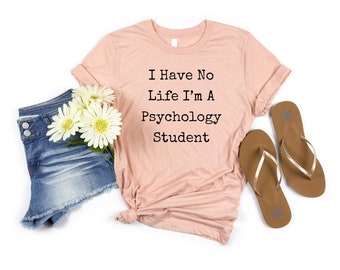 May include: A light pink short-sleeved t-shirt with the black text I Have No Life I'm A Psychology Student, paired with blue denim shorts and brown flip-flops, accented by a small cluster of white daisy flowers with yellow centers.
