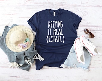May include: A navy blue t-shirt with the white text "KEEPING IT REAL (ESTATE)" printed on the front. The shirt is displayed alongside a straw hat with pastel flowers, a blue and white patterned scarf, white canvas sneakers, and brown sunglasses.