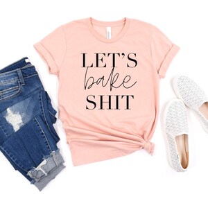 May include: A peach-colored short-sleeve t-shirt with the black text "LET'S bake SHIT" printed on the front, paired with dark blue distressed denim jeans and a pair of white slip-on shoes with a subtle grey textured pattern.