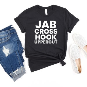May include: A black short-sleeve t-shirt with white text that reads JAB CROSS HOOK UPPERCUT, paired with blue denim jeans featuring distressed detailing and a pair of white slip-on shoes with a subtle textured pattern on the fabric surface.