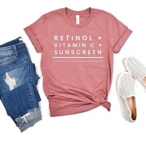 May include: A dusty rose t-shirt with rolled sleeves, featuring the text "RETINOL + VITAMIN C + SUNSCREEN" in white. The shirt is paired with blue jeans and white slip-on shoes with black stripes. The text below reads "(THESE ARE A FEW OF MY FAVORITE THINGS)".