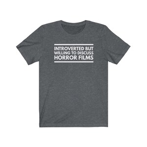 Może przedstawiać: Ciemnoszary melanżowy t-shirt z kr&oacute;tkim rękawem z białym napisem na środku klatki piersiowej: INTROVERTED BUT WILLING TO DISCUSS HORROR FILMS. Tekst jest umieszczony pomiędzy dwiema poziomymi białymi liniami, tworząc przejrzysty projekt graficzny.