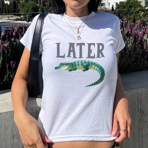 May include: White t-shirt with the word "LATER" printed in gray and a green alligator graphic.