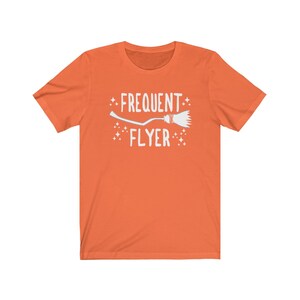 May include: Orange short-sleeved t-shirt with a white graphic of a broom and the text "Frequent Flyer".
