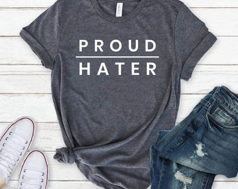 May include: A dark heather gray short-sleeve t-shirt featuring the white text PROUD HATER separated by a horizontal line. The shirt is displayed alongside a pair of blue distressed denim jeans and a pair of white canvas sneakers with white laces.