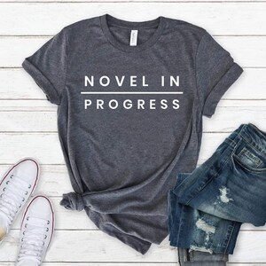 May include: A dark heather gray short-sleeve t-shirt featuring the white text NOVEL IN PROGRESS with a horizontal line separating the two words. The shirt has a crew neckline and is displayed alongside a pair of blue denim jeans and white sneakers.