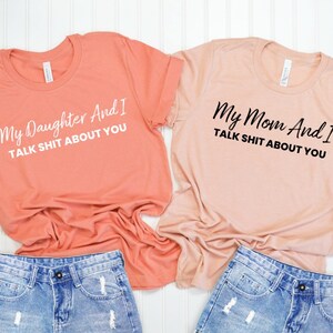 May include: Two peach colored t-shirts with a funny saying on each. The top shirt says "My Daughter And I TALK SHIT ABOUT YOU" in white text. The bottom shirt says "My Mom And I TALK SHIT ABOUT YOU" in black text.