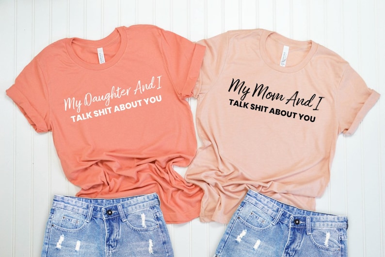 May include: Two peach colored t-shirts with a funny saying on each. The top shirt says "My Daughter And I TALK SHIT ABOUT YOU" in white text. The bottom shirt says "My Mom And I TALK SHIT ABOUT YOU" in black text.