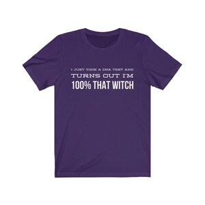 May include: A purple short-sleeved t-shirt featuring white text printed on the chest that reads: I JUST TOOK A DNA TEST AND TURNS OUT I'M 100% THAT WITCH. The garment has a crew neckline and a standard fit, showcasing a solid color design.