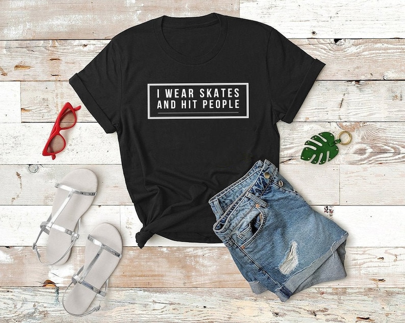 K&ouml;nnte beinhalten: Schwarzes T-Shirt mit wei&szlig;em Text, der "I wear skates and hit people" lautet. Das Shirt liegt auf einer wei&szlig;en Holzoberfl&auml;che mit einem Paar silbernen Sandalen, einer roten Sonnenbrille und einer blauen Jeans-Shorts.