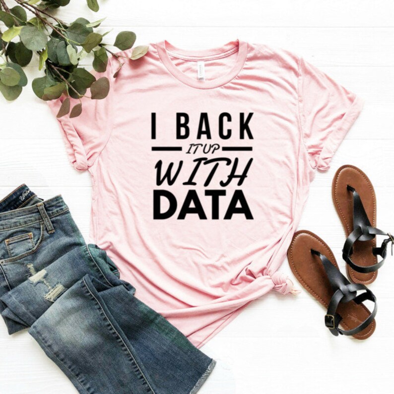 Puede incluir: Una camiseta de manga corta de color rosa claro con el texto negro I BACK IT UP WITH DATA impreso en la parte delantera. La camiseta se muestra junto a unos vaqueros azules y dos sandalias negras de tiras sobre una superficie de madera blanca.