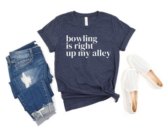 May include: A dark heather navy blue short-sleeve t-shirt with the white text "bowling is right up my alley" printed on the front, paired with distressed blue denim jeans and a pair of white slip-on shoes with a subtle textured pattern.