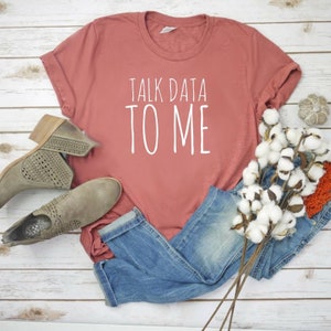 May include: A rust-colored t-shirt with the text "TALK DATA TO ME" in white lettering. The shirt is laid out on a white wooden surface with a pair of brown ankle boots and a bunch of white cotton flowers.