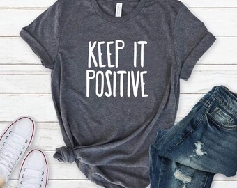 May include: A dark heather gray short-sleeve t-shirt featuring the white text "KEEP IT POSITIVE" printed in a clean, sans-serif font on the chest, paired with a pair of blue denim jeans and white canvas sneakers with white laces.