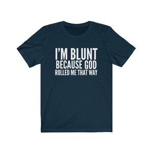 May include: Navy blue t-shirt with the white text "I'M BLUNT BECAUSE GOD ROLLED ME THAT WAY." The shirt has short sleeves and a crew neck. The text is centered on the front of the shirt.