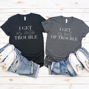 May include: A black t-shirt with the white text I GET us into TROUBLE and a dark grey t-shirt with the white text I GET us out OF TROUBLE, each paired with blue denim jeans and white canvas trainers with red and blue trim on the rubber soles.