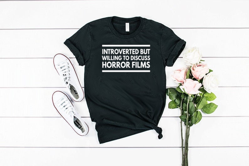 Może przedstawiać: Czarny t-shirt z kr&oacute;tkim rękawem z białym napisem "INTROVERTED BUT WILLING TO DISCUSS HORROR FILMS" na klatce piersiowej. Koszulka ma okrągły dekolt i jest pokazana obok białych trampek oraz bukietu r&oacute;żowych r&oacute;ż.