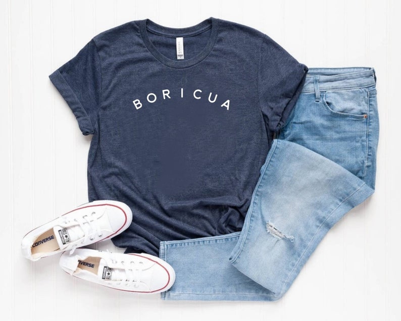 Puede incluir: Una camiseta azul marino con la palabra "BORICUA" en blanco, combinada con vaqueros de mezclilla azul claro y zapatillas de lona blancas. Las zapatillas tienen detalles rojos y se ve la palabra "CONVERSE".