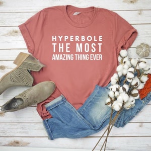 May include: A dusty rose short-sleeved t-shirt with white text that reads HYPERBOLE THE MOST AMAZING THING EVER. The shirt is paired with blue denim jeans, a pair of taupe perforated ankle boots, and a decorative stem of white cotton bolls.