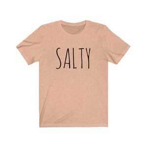 May include: A light peach colored t-shirt with the word "SALTY" printed in black on the front.