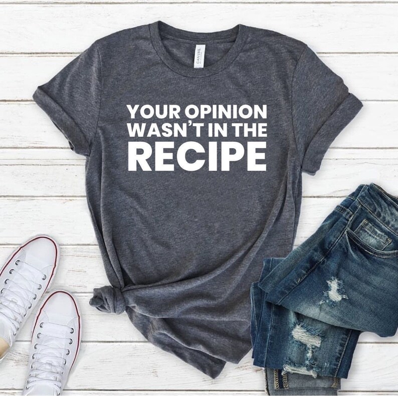 May include: A dark heather grey short-sleeve t-shirt featuring the white text "YOUR OPINION WASN'T IN THE RECIPE" printed on the front. The shirt is displayed alongside a pair of blue denim jeans and white canvas trainers on a white wooden surface.