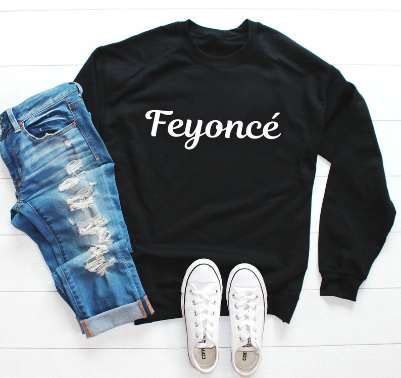 K&ouml;nnte beinhalten: Schwarzer Pullover mit wei&szlig;em Text, der "Feyonc&eacute;" auf wei&szlig;em Hintergrund sagt. Der Pullover wird mit einer blauen Jeans und wei&szlig;en Turnschuhen kombiniert.