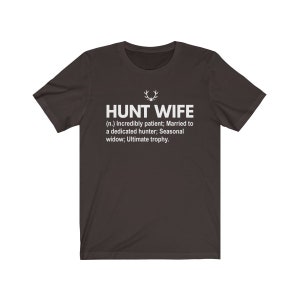 May include: A dark gray t-shirt with white text that reads "HUNT WIFE (n.) Incredibly patient; Married to a dedicated hunter; Seasonal widow; Ultimate trophy."