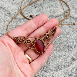Op de afbeelding: Een ketting met een rode carneol edelsteen hanger, een bruin koord en koperen kralen. De hanger is in een geknoopt macramé ontwerp.