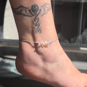 Op de afbeelding: Een zilveren enkelbandje met drie iriserende opaalstenen op een grijs koord.