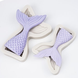May include: Two silicone moulds for baking mermaid tails. The moulds are in the shape of a mermaid tail with scales and fins. The moulds are light purple and white.