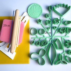 May include: A set of green plastic cookie cutters in various shapes, including flowers, leaves, and a fan. The set also includes a rolling pin, two wooden dowels, a pink sponge, and a white plastic tool. The set measures 20 cm by 26 cm.