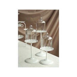Puede incluir: Tres soportes para tartas de metal blanco con cúpulas de vidrio transparente. Cada soporte tiene una base redonda y una sola varilla de soporte vertical y fina. Las cúpulas tienen un borde festoneado.