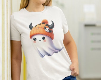 May include: A white short-sleeved t-shirt featuring a graphic of a cute white ghost wearing an orange knit beanie with black horns and a small ghost pin. The ghost has large black eyes, rosy cheeks, and a small mouth, rendered in a cartoon style.