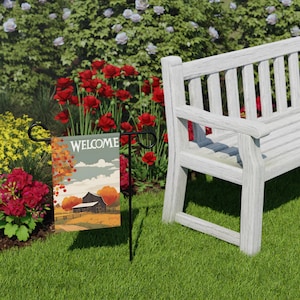 Puede incluir: Una bandera de jardín con la palabra "WELCOME" en blanco. La bandera presenta una escena otoñal con un granero, árboles y una valla. La bandera está en un soporte de metal negro en un jardín con un banco blanco y flores.