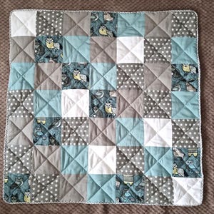 Könnte beinhalten: Ein Patchwork-Quilt mit Quadraten aus grauem, weißem und blaugrünem Stoff. Einige Quadrate haben ein Sternmuster, während andere Fuchs- und Eulenmuster aufweisen. Der Quilt ist etwa 76 cm groß.