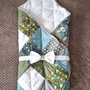 Könnte beinhalten: Eine Baby-Swaddle-Decke mit Patchwork-Design. Die Decke hat eine weiße Schleife und ein gemustertes Design mit grünen, blauen und grauen Quadraten. Die Decke ist gesteppt und hat eine weiße Innenseite.