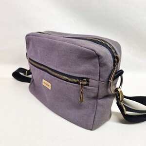 Könnte beinhalten: Eine graue Canvas-Umhängetasche mit schwarzem Reißverschluss und Riemen. Die Tasche hat eine kleine Tasche auf der Vorderseite mit einem braunen Leder-Etikett, auf dem "The Remarkable" steht.